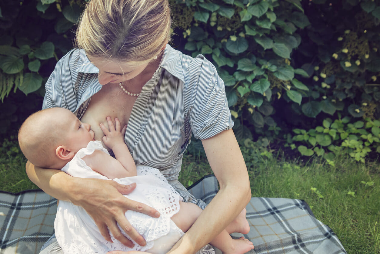 Mother breastfeeding baby girl outdoors in summer nature. Mother is wearing a classic striped cotton dress and a pearl necklace, baby girl is 4 month and in a white dress. Both have very fair skin. They are sitting on a plaid on the grass. Horizontal waist up shot with copy space.