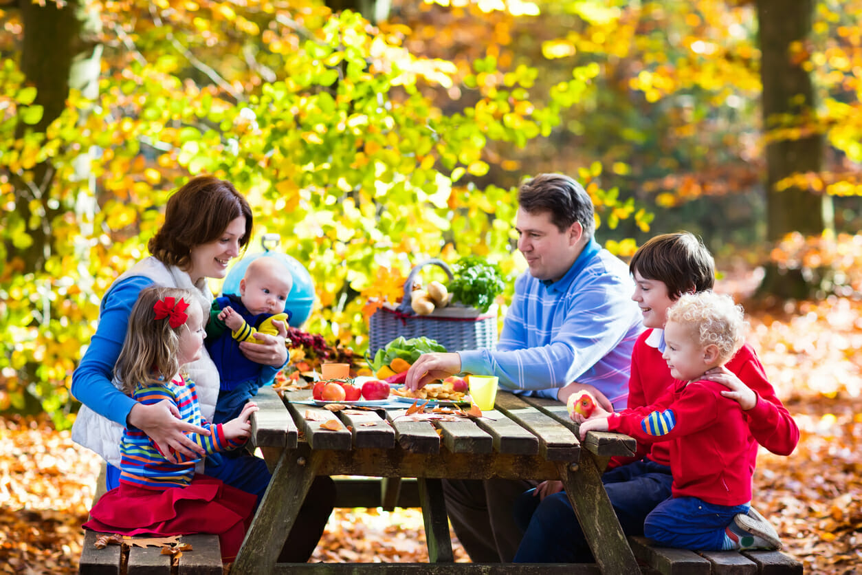 Happy young family with four children grilling meat and making sandwich and salad on a picnic table in sunny autumn park. Barbeque fun for parents with kids on warm fall day. Grill and BBQ party.